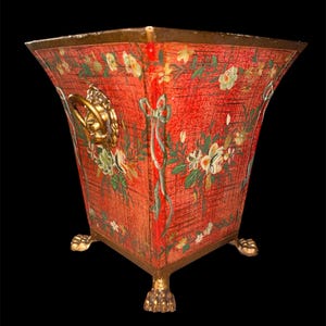 Puede incluir: Una jardinera roja y cuadrada con diseños florales y detalles dorados. La jardinera tiene una superficie texturizada, un borde dorado y asas de cabeza de león doradas. Descansa sobre patas de garra doradas.