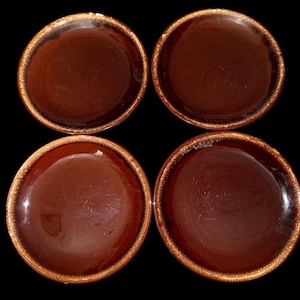 May include: A set of four round ceramic saucers featuring a glossy dark brown center and a textured, lighter brown rim. The plates are arranged in a two-by-two grid pattern against a solid black background, showcasing a smooth, reflective surface.