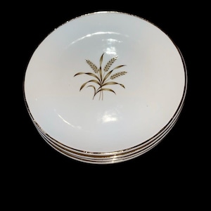 May include: A stack of four white ceramic plates with a gold rim and a wheat stalk design in the center. The wheat design is in shades of brown and gold. The plates are stacked on top of each other.