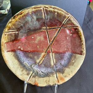 May include: A round ceramic plate with a brown, red, and white glaze. The plate has a unique design with a series of lines that create a geometric pattern.