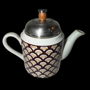 May include: A white ceramic teapot featuring a repeating brown, cream, and gold fan-shaped pattern on the body. The teapot includes a white handle and spout, topped with a metallic silver lid that has a small, rounded wooden knob at the center.