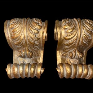 Puede incluir: Dos ménsulas arquitectónicas antiguas de color dorado con diseños ornamentados de hojas y volutas. Las ménsulas tienen un aspecto envejecido, con una superficie texturizada y un fondo oscuro. Cada ménsula tiene una base decorativa y redondeada.