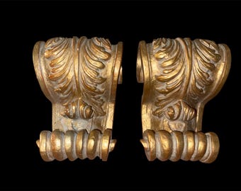 May include: Two antique gold-colored architectural corbels with ornate leaf and scroll designs. The corbels have a weathered appearance, with a textured surface and a dark background. Each corbel has a decorative, rounded base.