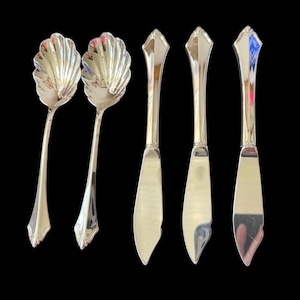 May include: A set of five silver-toned metal utensils consisting of two spoons with scalloped, shell-shaped bowls and three knives with pointed, tapered blades. Each piece features a smooth, reflective finish and a decorative, angular handle design.
