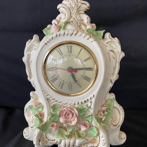 May include: A white ceramic mantel clock with a gold-tone clock face and Roman numerals. The clock is adorned with pink roses and green leaves, with gold accents. The clock has a decorative, ornate design and stands on three feet.