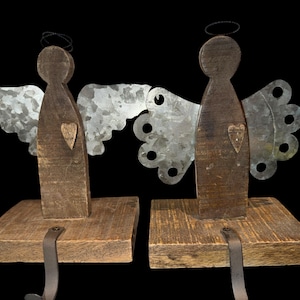 May include: Two angel-shaped stocking holders made of dark brown wood with galvanized metal wings. Each figure features a small wooden heart on the chest, a thin wire halo above the head, and a metal hook attached to the square wooden base.