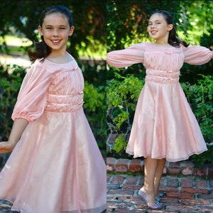 May include: A young girl wearing a pale pink dress with long sleeves and a gathered bodice. The dress has a full skirt and is made of a sheer fabric. The girl is standing in a garden setting.