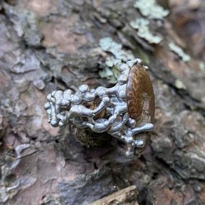 Pu&ograve; includere: Un anello unico con una pietra marrone ovale con un motivo strutturato, incastonato in una fascia di metallo argentato dall'aspetto organico. L'anello &egrave; esposto su una superficie marrone strutturata, forse legno o corteccia.