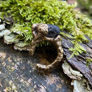Puede incluir: Un anillo de color bronce con un diseño orgánico rugoso, con una piedra oscura y texturizada. El anillo se encuentra sobre un fondo de musgo y madera, resaltando su estética natural. Su forma y color únicos lo convierten en una joya distintiva.