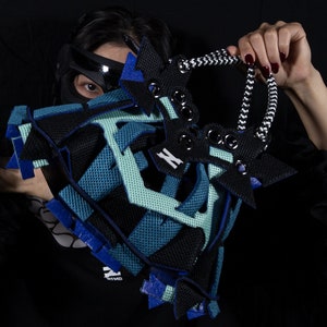 May include: A black, blue, and green geometrically shaped bag with a black and white rope handle. The bag is made of a mesh material and has a variety of different textures.