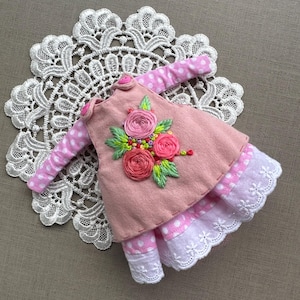 May include: A miniature pink dress with white polka dot sleeves and a white lace hem. The dress is adorned with embroidered pink roses and green leaves. The dress is displayed on a white lace doily.