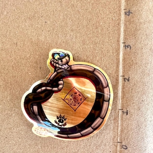 May include: A colorful sticker featuring a cartoon snake with a smiling face and blue eyes. The snake is brown and black, coiled around a wooden-textured center with a paw print and spiral design.