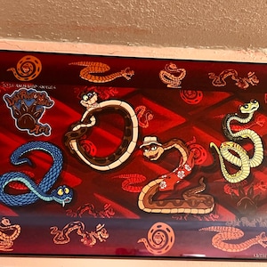 Puede incluir: Una impresión artística rectangular enmarcada que muestra el año 2025 formado por serpientes de dibujos animados sobre un fondo rojo oscuro. Los números están formados por una serpiente marrón, una con sombrero y camisa, y una serpiente amarilla.