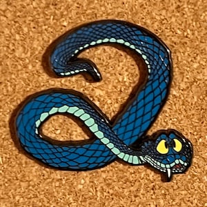 May include: A blue snake-shaped enamel pin with a detailed scale pattern. The pin features a light blue stripe along its back and a smiling face with yellow eyes. The pin is set against a textured, brown background.