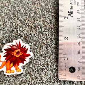 May include: A sticker of a cartoon lion with a red mane roaring. The lion is orange and has a paw print on its tail. The sticker is about 2 inches tall.