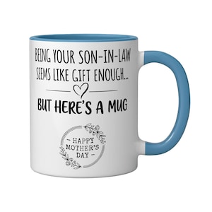 Op de afbeelding: Witte keramische mok met een blauw handvat en rand. De mok heeft de tekst "BEING YOUR SON-IN-LAW SEEMS LIKE GIFT ENOUGH... BUT HERE'S A MUG" en "HAPPY MOTHER'S DAY" in zwarte letters.