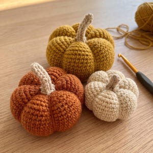 Op de afbeelding: Drie handgemaakte gehaakte pompoenen in mosterdgeel, oranje en crème. Elke pompoen heeft een textuur met ribbels en een kleine, lichtgekleurde gehaakte steel die in het midden bovenop het ronde, opgevulde lichaam is bevestigd.