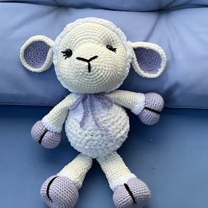 May include: A handmade crochet lamb plush toy featuring a cream-colored body and head with light purple accents on the ears, hooves, and a small bow tied around the neck. The lamb has embroidered black eyes and a nose, with a textured yarn finish.