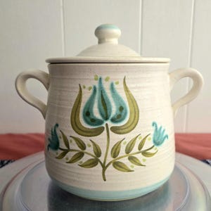 May include: Cream ceramic lidded pot with a floral design in teal and olive green. The pot has two handles and a teal band around the base. The lid has a small knob and a matching teal accent.