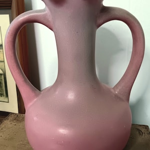 May include: A pink ceramic vase with two handles and a ruffled top. The vase has a gradient of color, fading from a lighter pink at the top to a darker pink at the bottom.