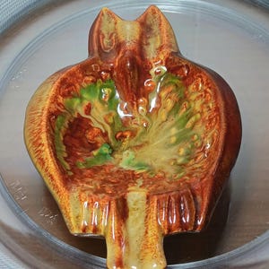 May include: A ceramic pomegranate-shaped dish with a glossy finish. The dish features a blend of orange, brown, and green hues, with a detailed interior design. The base is a light tan color, and the dish is set inside a clear, round container.