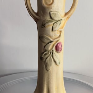 May include: A cream-colored ceramic vase with two handles and a flared base. The vase is decorated with embossed leaves and a pink fruit. The vase has a textured, crackled surface, and is approximately 8 inches tall.