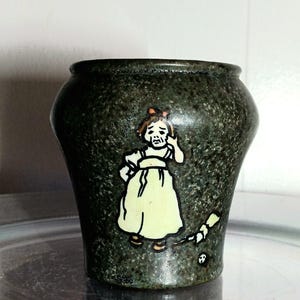 May include: A dark green speckled ceramic vase with a cartoon illustration of a girl in a yellow dress. The girl is holding her hand up to her head. A broom and a skull are also illustrated on the vase.