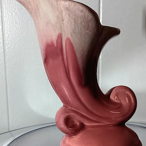 May include: A vintage ceramic vase in shades of pink, with a distinctive, curved form. The vase has a flared top, a swirling design, and a solid base. The colour transitions from light pink to a darker rose hue, creating a gradient effect.