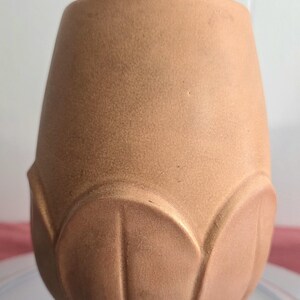May include: A matte, light brown ceramic vase with a smooth, tapered cylindrical body. The lower section features a decorative, raised petal-like relief pattern that wraps around the base, giving the vessel a distinct, sculpted appearance.