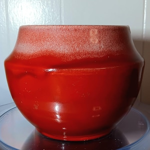 May include: A red ceramic planter with a white glaze on the top rim. The planter has a rounded shape and a smooth, glossy finish.