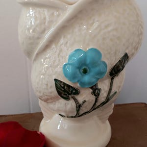 May include: A white ceramic vase with a textured surface and a blue flower design. The vase has a unique shape with a wide opening and a narrow base.