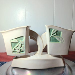 May include: Cream-colored ceramic planter with two connected compartments. Each compartment features a rectangular, light green inset with a leaf design. The planter has a curved support and a wide, flat base.