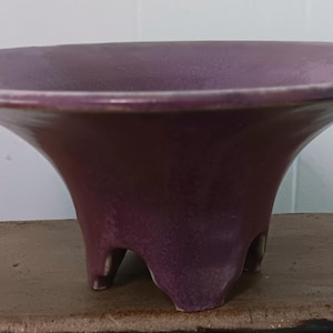 May include: A purple ceramic planter with a wide, shallow bowl and three legs. The planter has a subtle, mottled glaze.