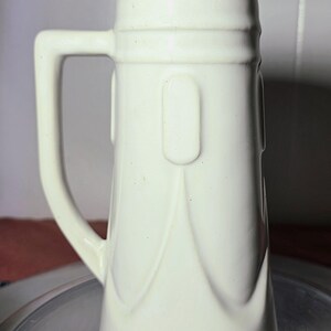 May include: A white ceramic beer stein with a handle. The stein has a rectangular shape with rounded edges and a decorative design. The handle is a sturdy, rectangular shape. The stein is empty and clean.