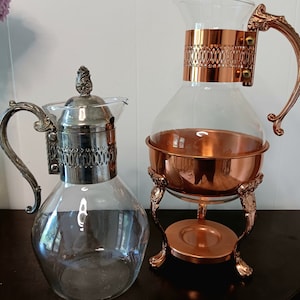 May include: Two glass coffee pots with copper accents. The pot on the left has a silver lid and base. The pot on the right has a copper base and stand with ornate feet.