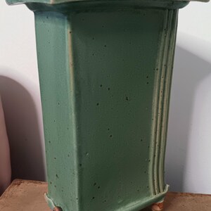 May include: A green ceramic vase with a rectangular body and a square top. The vase has a textured surface and four small feet.
