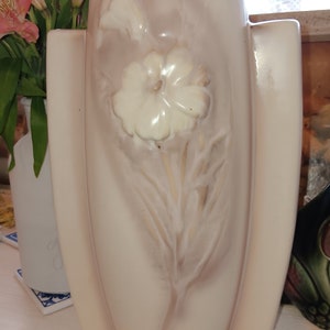 May include: A white ceramic vase with a raised floral design. The vase has a light pink glaze and features a single white flower with leaves.