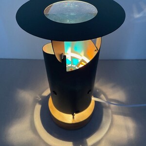 Könnte beinhalten: Eine schwarze Tischlampe mit einer runden, flachen Oberseite und einem zylindrischen Sockel. Die Lampe hat einen klaren Glasschirm und ein weißes Kabel.