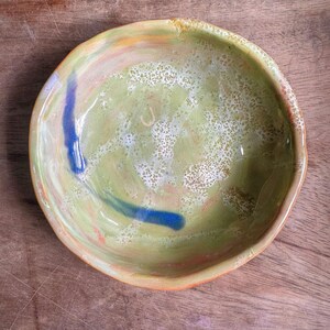 May include: Handmade ceramic bowl with an irregular shape. The interior is pale green with a textured surface, featuring blue and orange brushstrokes. The rim has a warm, earthy tone, suitable for serving or display.