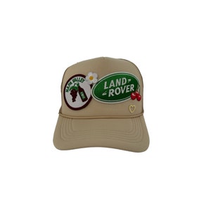 Peut inclure: Un chapeau de camionneur beige avec un patch "LAND-ROVER" vert et blanc, un patch de fleur blanche et un patch "NAPA VALLEY" vert et marron avec une bouteille de vin et des raisins.