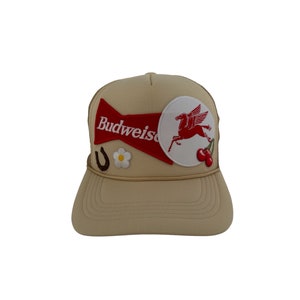 Peut inclure: Un chapeau de camionneur beige avec un logo Budweiser rouge, un patch blanc avec un cheval ailé rouge, un fer à cheval et une fleur blanche.