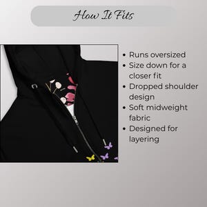 May include: Black zip-up hoodie with a floral and butterfly pattern on the inside lining. The hoodie has a drawstring hood and a silver zipper. The text "How It Fits" is at the top of the image.