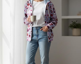May include: A light blue zip-up hoodie with a floral pattern in shades of red and purple. The hoodie has a light blue lining and a drawstring. The person is wearing a white t-shirt and light blue jeans, holding a beige mug.