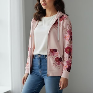 May include: A pale pink zip-up hoodie featuring a floral pattern in red, pink, and burgundy. The hoodie has a drawstring hood, front pockets, and a white zip. The model is wearing a white t-shirt and blue jeans.