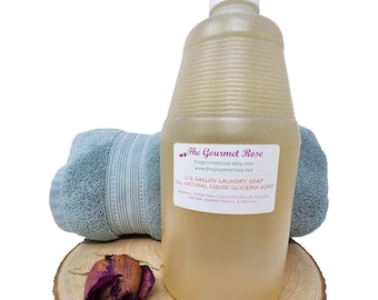 Puede incluir: Una botella grande y transparente de jabón líquido para ropa con tapa blanca y etiqueta. La botella está sobre una rodaja de madera con una rosa seca y una toalla azul doblada. La etiqueta dice "The Gourmet Rose".