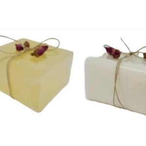 May include: Two square soaps, one pale yellow and one white, are tied with twine and adorned with dried rosebuds. The soaps have a rustic, handmade appearance, suggesting a natural skincare product.