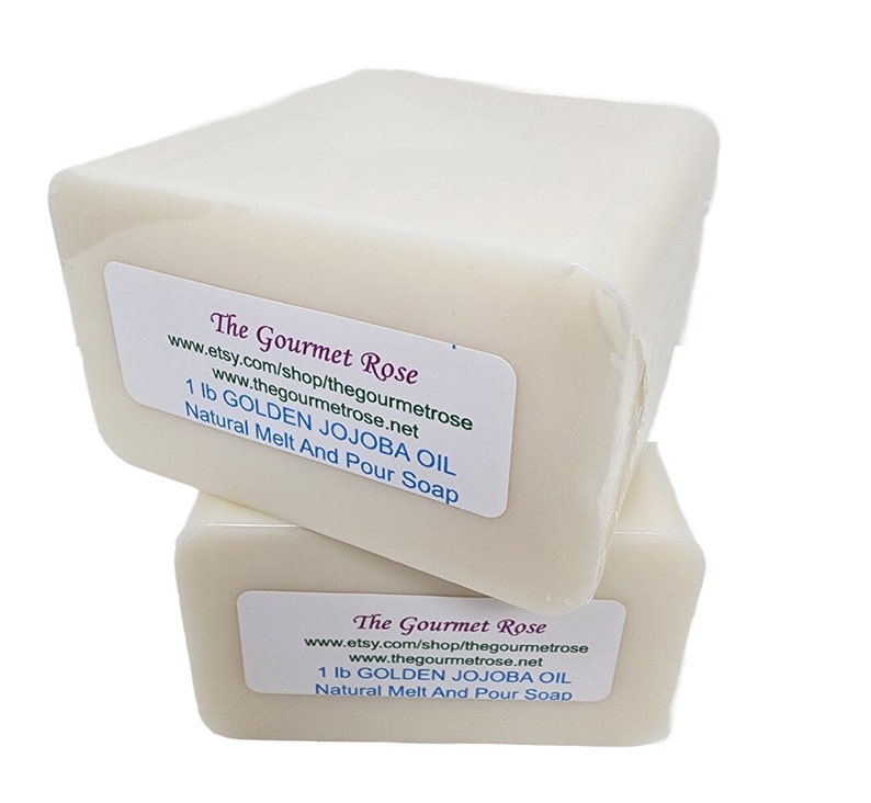 Op de afbeelding: Twee ivoorkleurige rechthoekige zeepblokken op elkaar gestapeld. Elk blok heeft een label met de tekst "The Gourmet Rose", websiteadressen en "1 lb GOLDEN JOJOBA OIL Natural Melt And Pour Soap".
