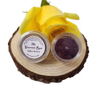 Può includere: Due piccoli vasetti di ombretto su una fetta di legno, accanto a una rosa gialla. Un vasetto contiene ombretto marrone, etichettato "Sultry Brown" da "The Gourmet Rose". L'altro vasetto contiene ombretto viola scuro. Ingredienti elencati.