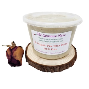 May include: A clear plastic container of organic raw shea butter, labeled "The Gourmet Rose," sits on a small wooden coaster. The label indicates the product is 100% pure and weighs 1 lb. A dried rose bud rests to the left of the container.