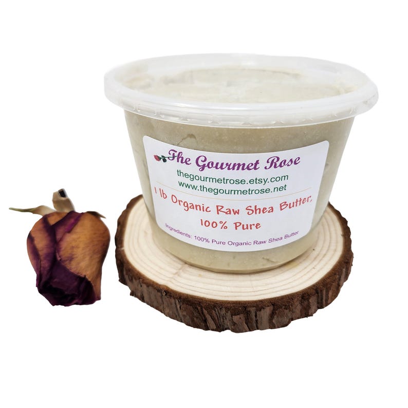 May include: A clear plastic container of organic raw shea butter, labeled "The Gourmet Rose," sits on a small wooden coaster. The label indicates the product is 100% pure and weighs 1 lb. A dried rose bud rests to the left of the container.